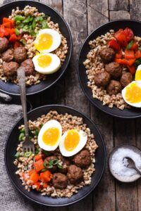 Overhead view of three grain bowls topped with sausage meatballs, fresh vegetables, and boiled egg.