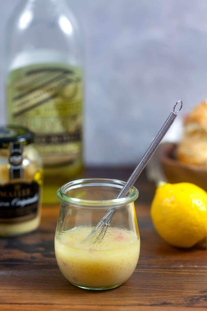 A small jam jar of homemade lemon vinairgette on a wooden surface, with the ingredients in the background. 