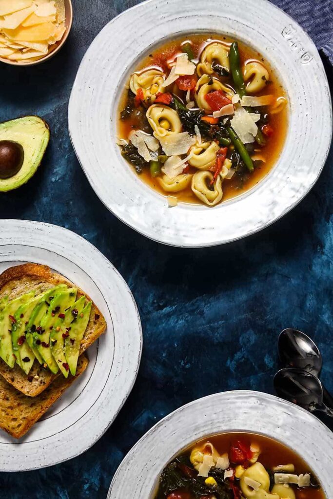 Two bowls of tortellini soup and a plate of avocado toast on a blue background.