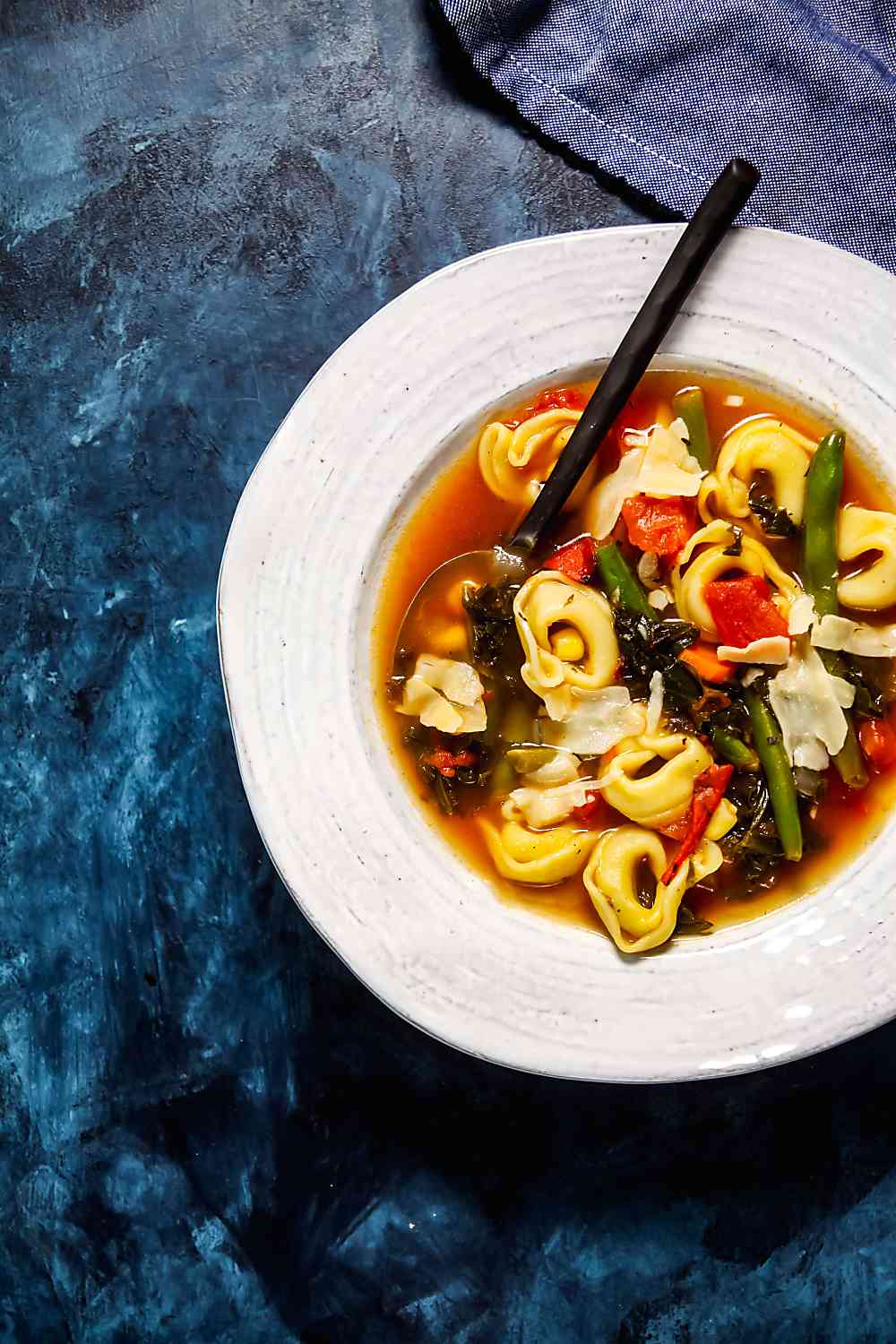 Overhead view of a bowl of tortellini vegetable soup on a blue background. 