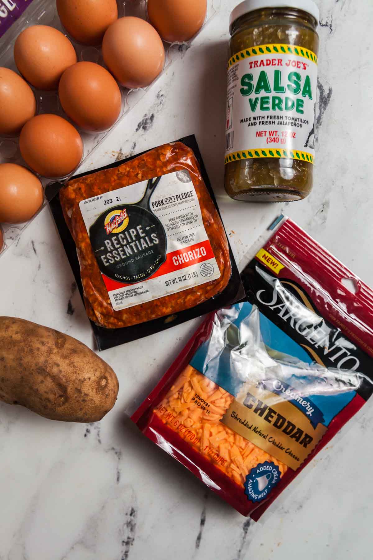 Overhead image of ingredients needed to make a chorizo and potato breakfast burrito on a white marble background. Ingredients include eggs, green salsa, chorizo, cheese, and a potato. 