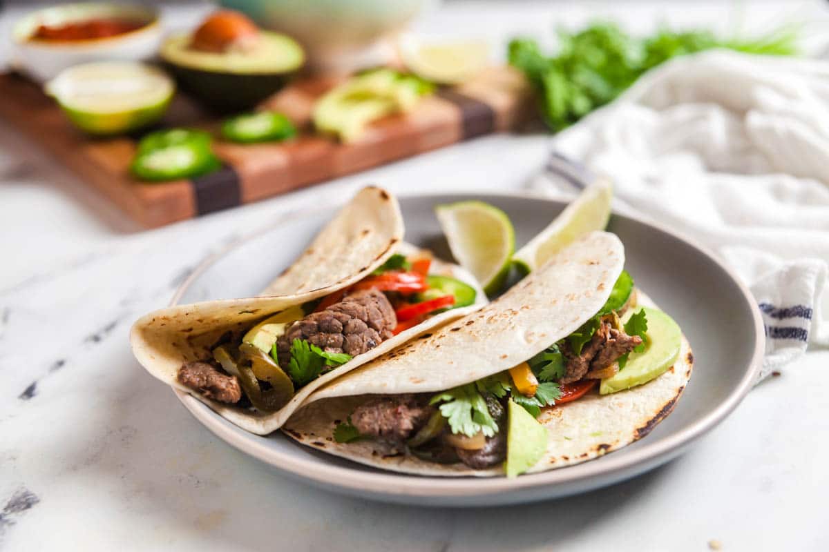Landscape image of two steak fajitas on a plate, with a wooden board of topping options in the background.