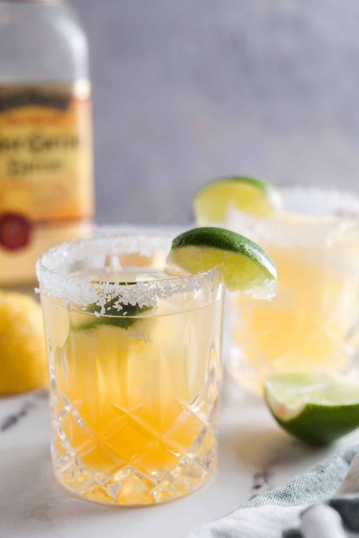 Two margaritas with salted rims on a marble table, with a bottle of tequila in the background.