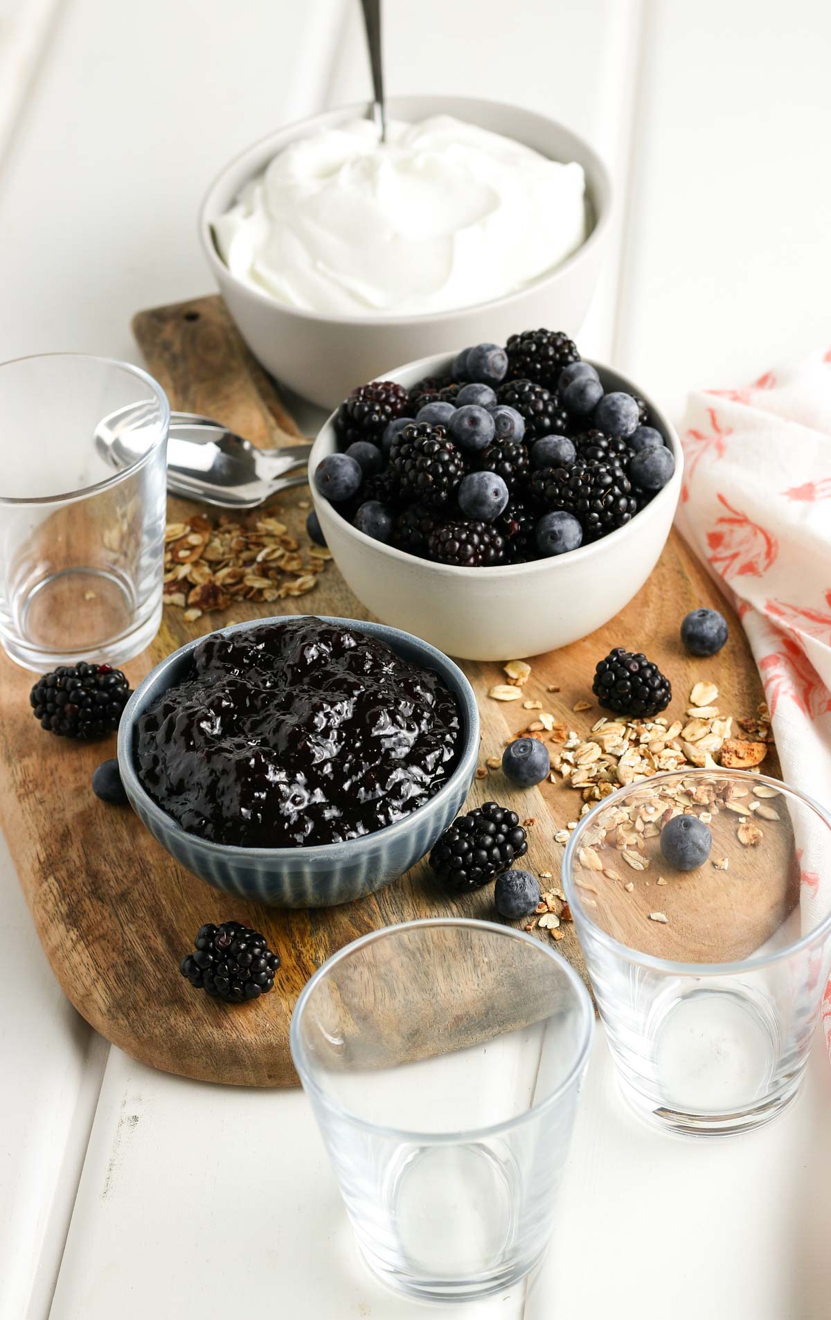 A wooden board with the ingredients needed to make a blackberry yogurt parfait, including Greek yogurt, blackberry jam, fresh berries, and granola. 