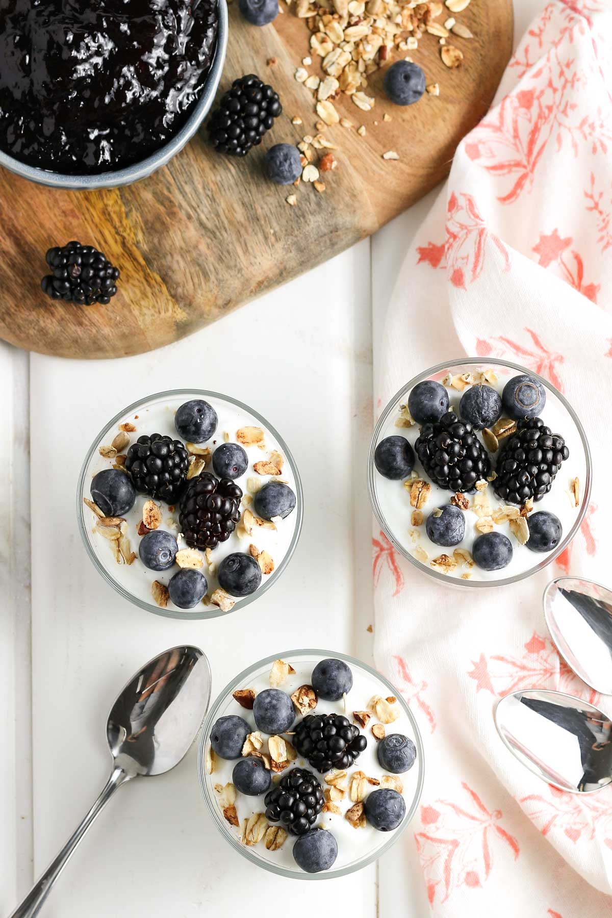 An overhead view of three blackberry yogurt parfaits with ingredients in the background. 