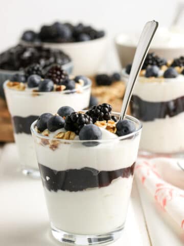 Layered Greek yogurt parfaits with blackberries, blueberries, and granola in individual glasses. The blackberry parfait in the front has a spoon in it.