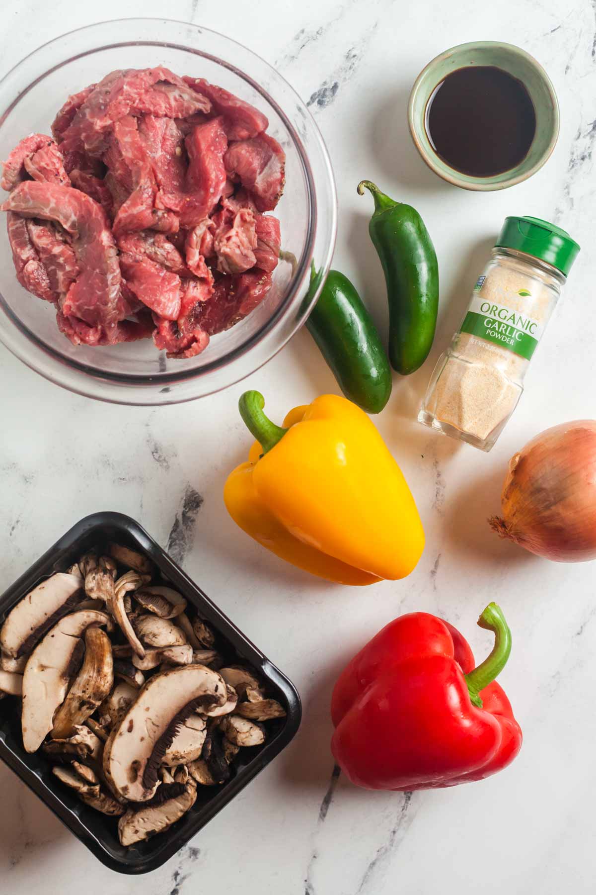 Ingredients for making easy Philly cheesesteak sandwiches, including sliced steak, peppers, onion, mushrooms, jalapeno peppers, Worchestershire sauce, and garlic powder.