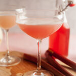 A rhubarb gin sour cocktail on a wooden board with a bottle of rhubarb syrup in the background.