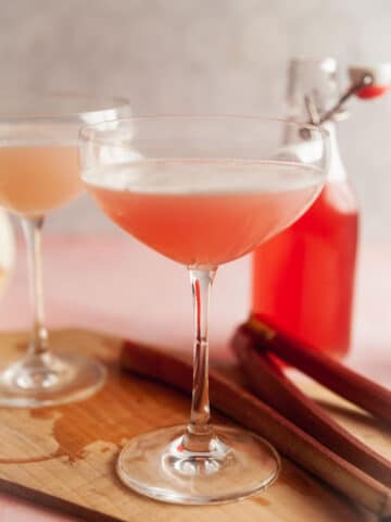 A rhubarb gin sour cocktail on a wooden board with a bottle of rhubarb syrup in the background.