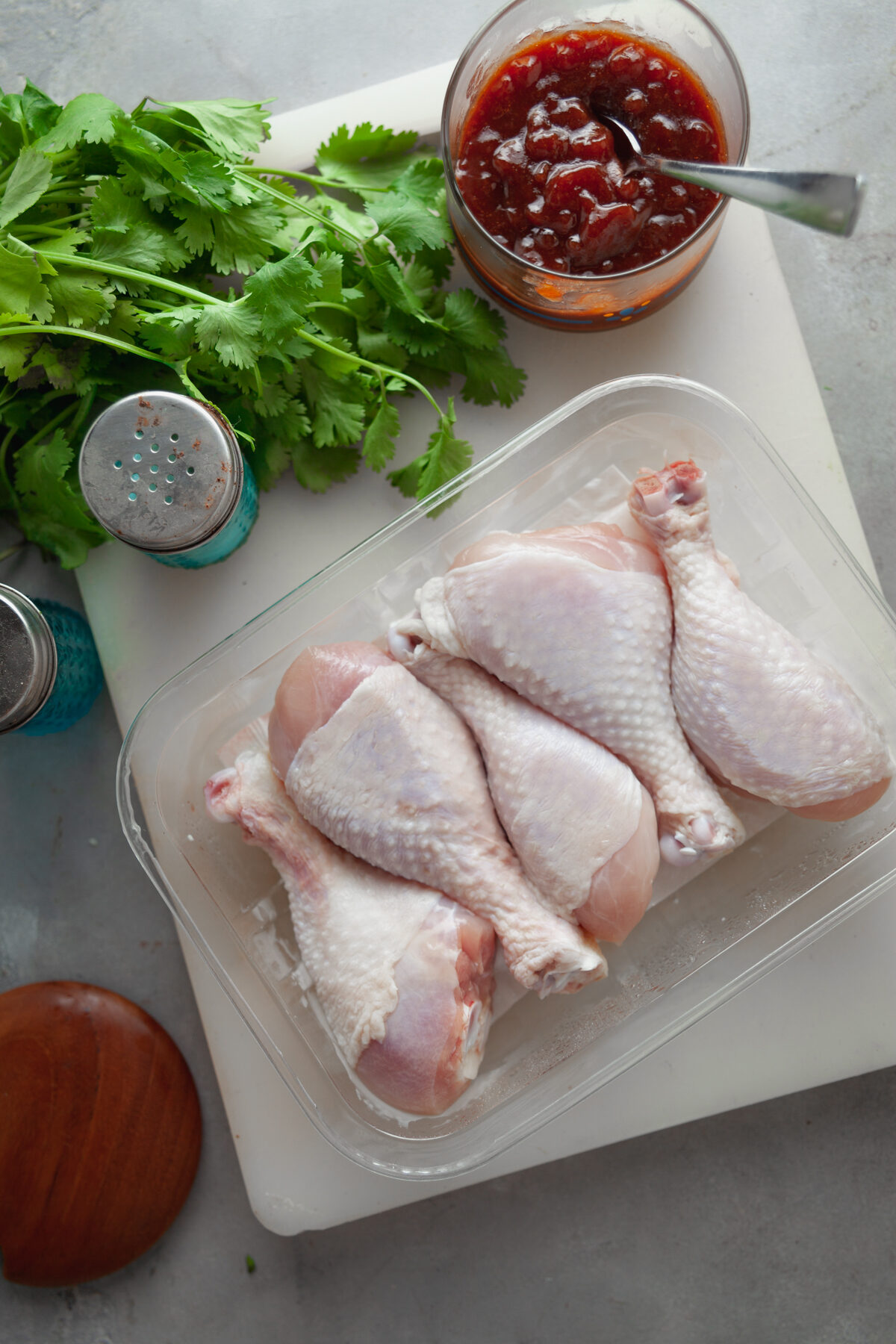 Overhead view of raw chicken legs with salt and pepper shakers, parsley, and a container of bbq sauce. 