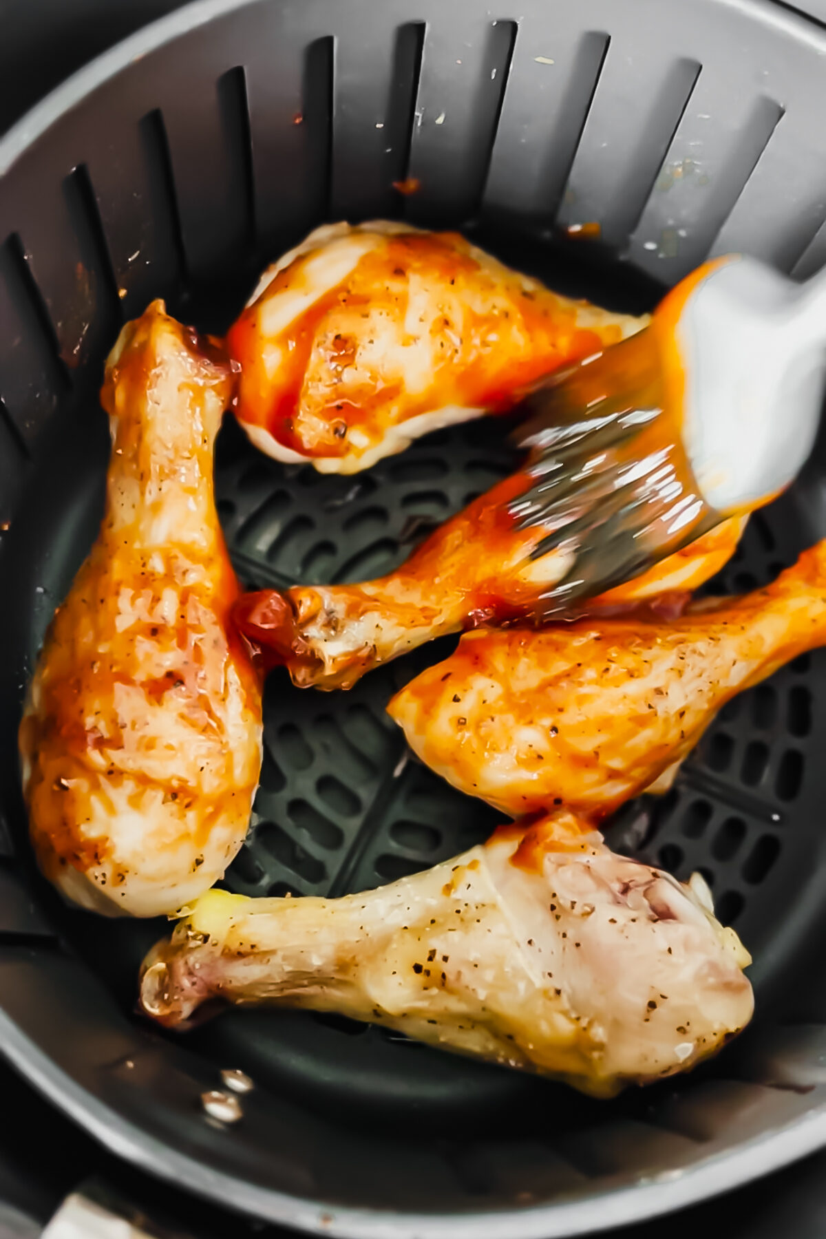 Brushing bbq sauce on chicken drumsticks in the air fryer basket. 