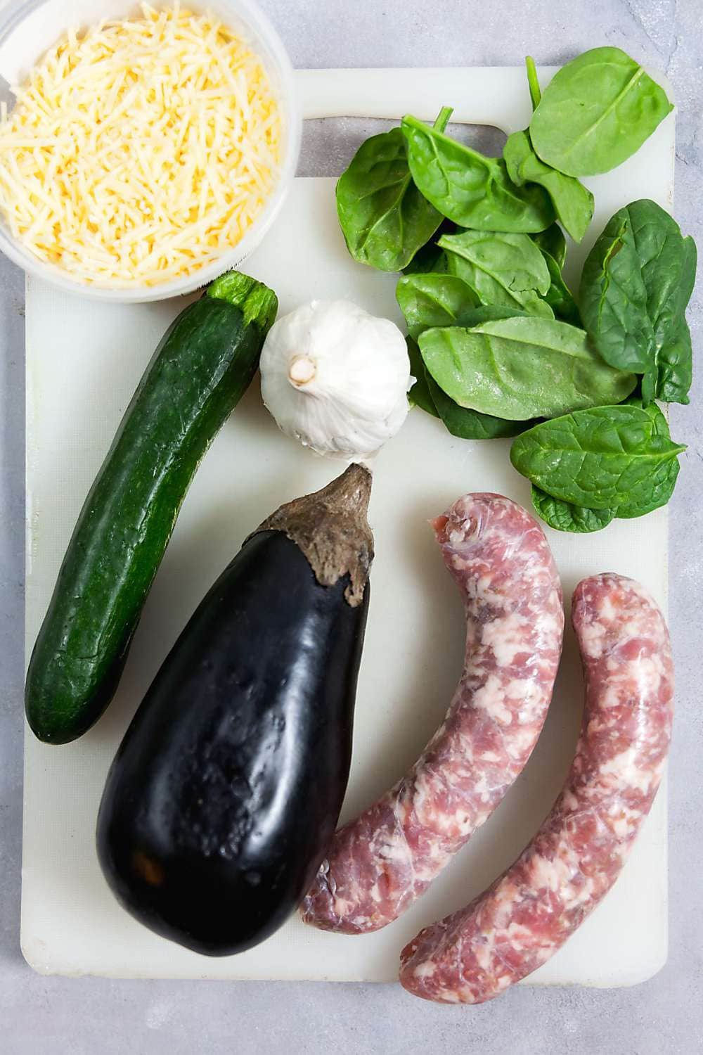 Sausage, eggplant, garlic, spinach, and parmesan cheese shreds on a white cutting board.