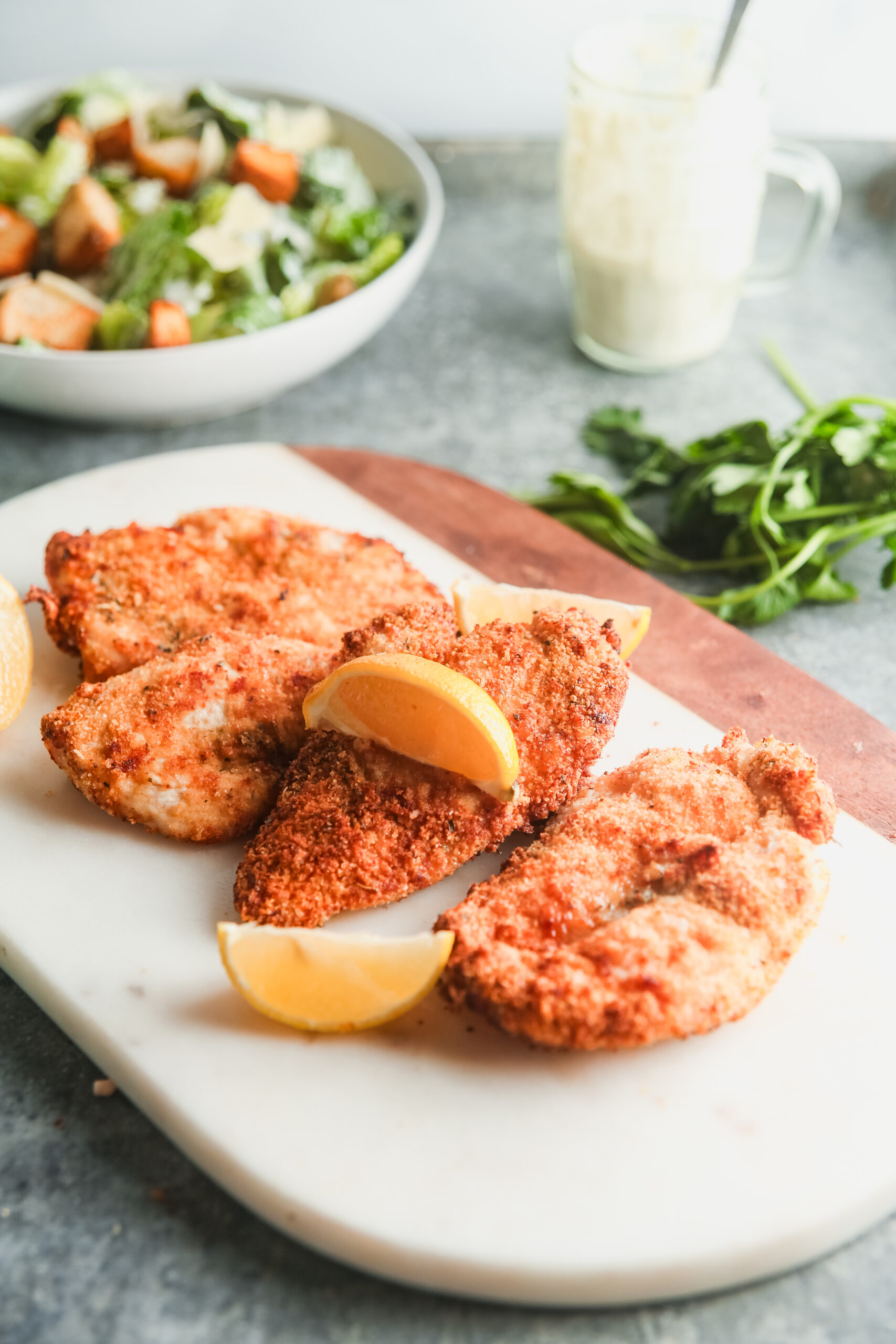 A pile of air fryer chicken cutlets and lemon wedges on a white serving board, with a salad in the background.