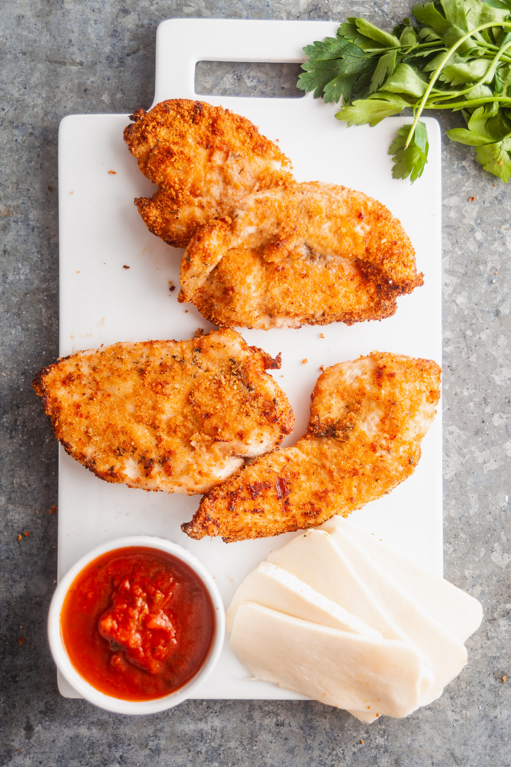 Prepping chicken parmesan: breaded chicken cutlets, marinara sauce, and mozzarella cheese on a white platter.