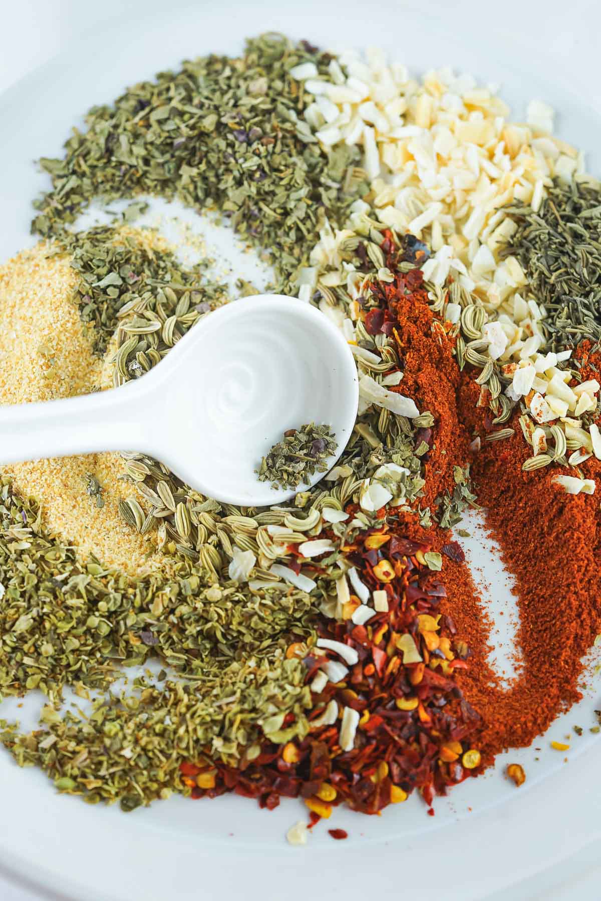 A small dish filled with dried herbs needed to make a custom seasoning blend, with a spoon swirling them together.