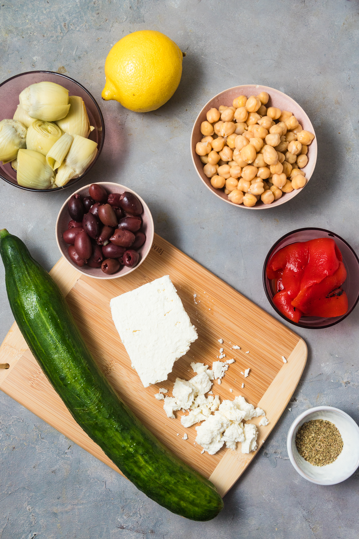 Ingredients needed to make Mediterranean chickpea salad with artichokes and cucumber arranged on a slate surface. 