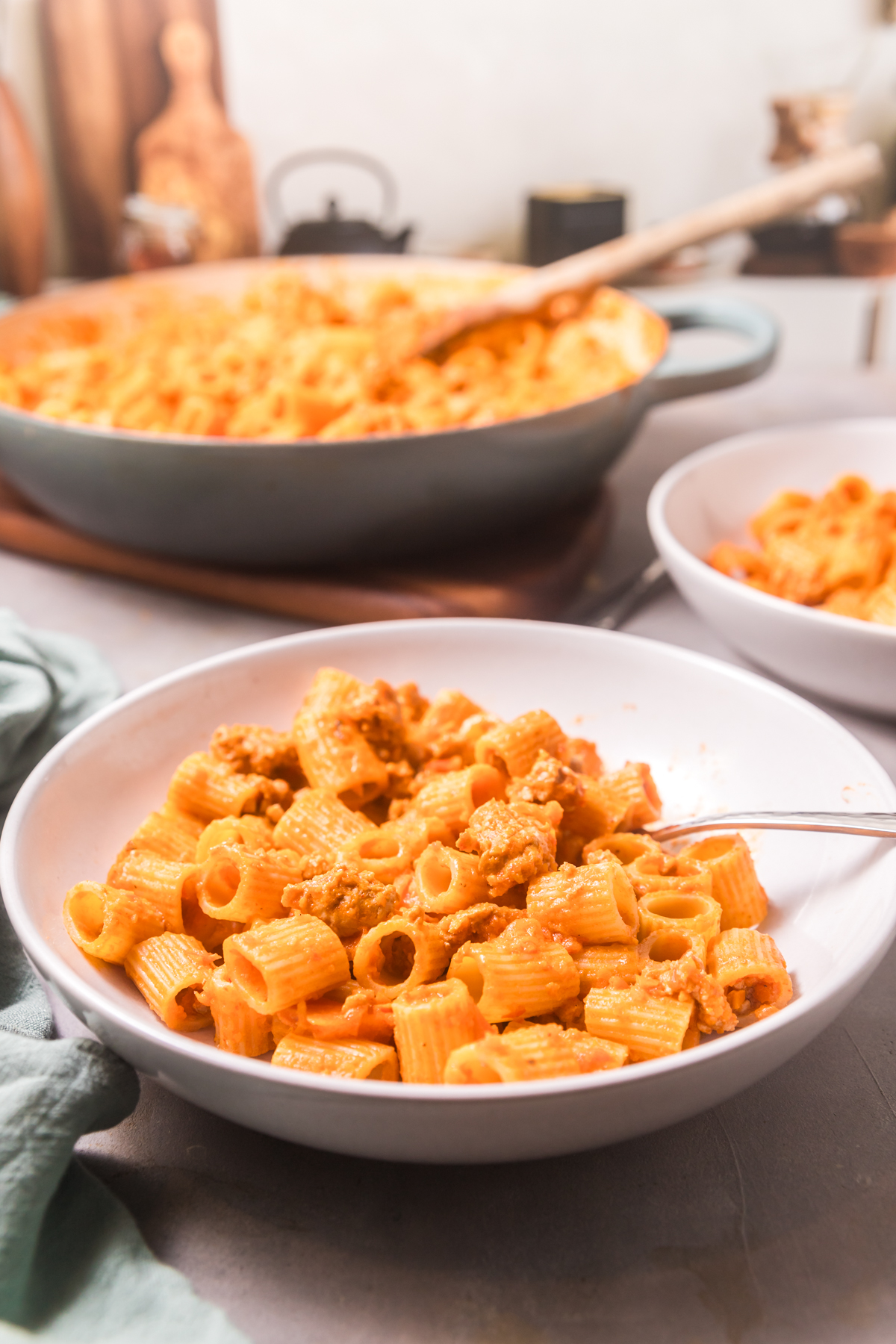 A dining scene with the focus on a white bowl of thai red curry pasta and a pan of pasta in the background. 