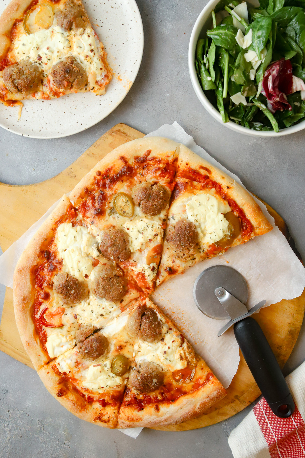 Overhead view of a homemade meatball pizza with a pizza wheel and a side salad. 