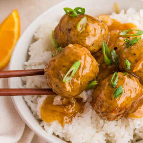A bowl of rice topped with orange glazed meatballs being lifted by chopsticks.
